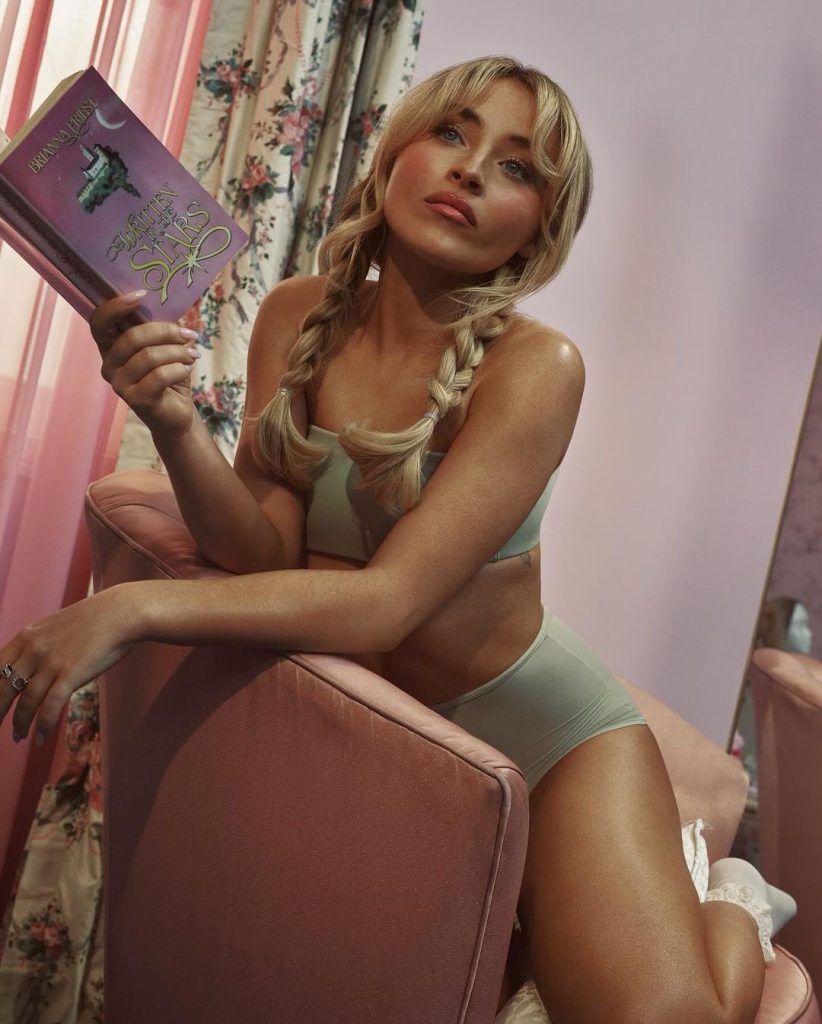 L'actrice Sabrina Carpenter pose pour Skims dans une tenue grise sportive, assise sur un fauteuil en cuir en tenant un livre.
